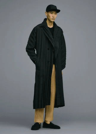 Asian male fashion model posing in a pleated black coat and pants, in a studio with grey background.