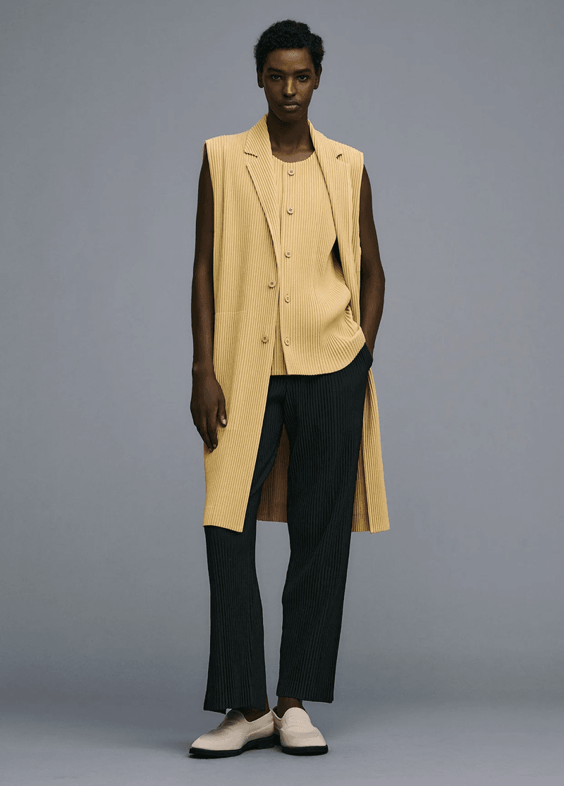 An adult female model poses in a photo studio wearing a long sleeveless yellow pleated vest, button-up shirt, wide pleated black pants, and shoes.