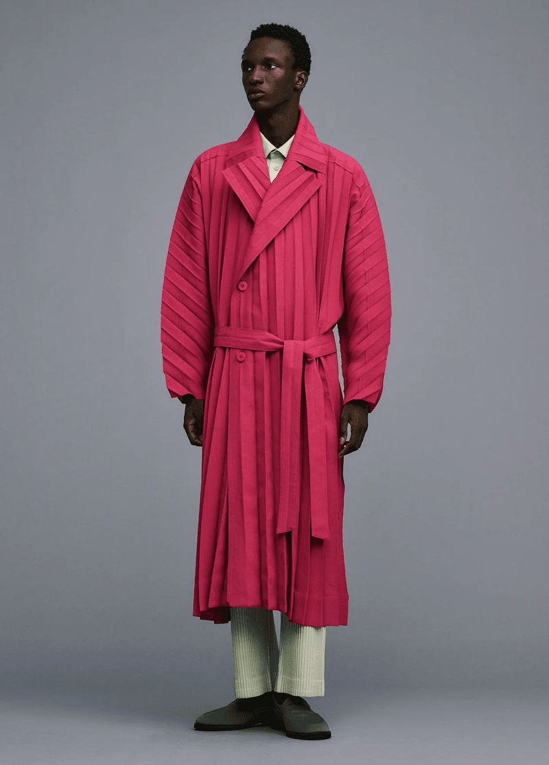 Dark-skinned male fashion model poses in a studio wearing a pink pleated trench coat with a belt, matching pants, and casual shoes.