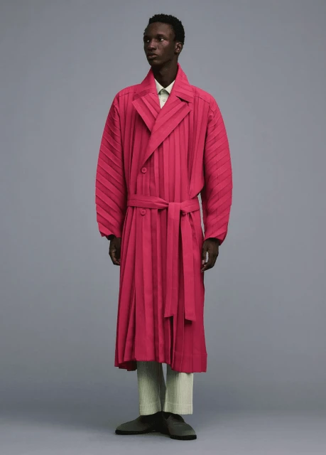 Dark-skinned male fashion model poses in a studio wearing a pink pleated trench coat with a belt, matching pants, and casual shoes.