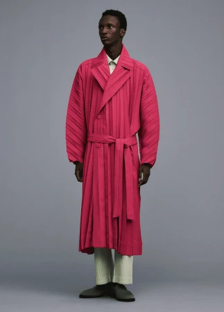 Dark-skinned male fashion model poses in a studio wearing a pink pleated trench coat with a belt, matching pants, and casual shoes.