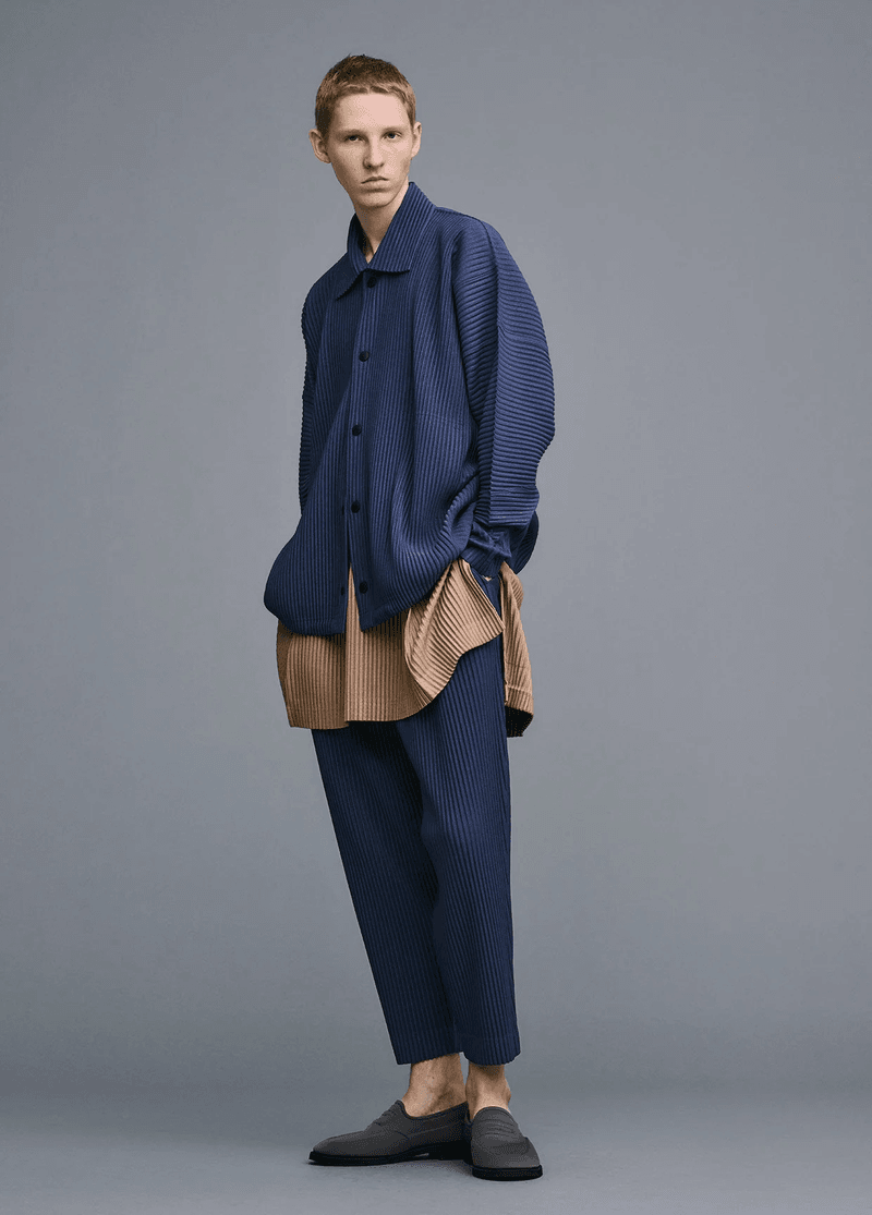 Male model in pleated blue shirt, tan top, pleated blue pants, and dark slip-on shoes in a studio setting with gray backdrop.