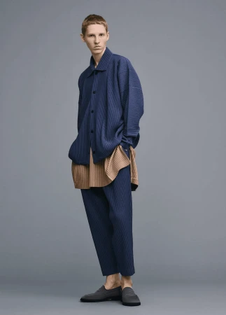 Male model in pleated blue shirt, tan top, pleated blue pants, and dark slip-on shoes in a studio setting with gray backdrop.
