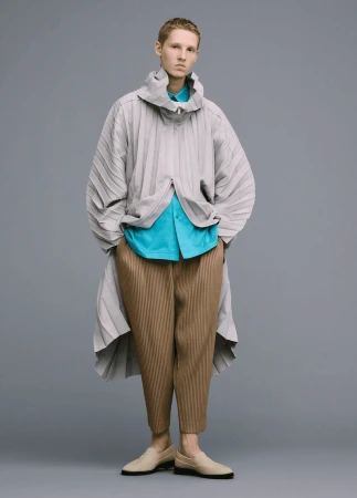 Full-length shot of an adult male model in a light grey pleated jacket and pants with a blue shirt, on a grey studio background.