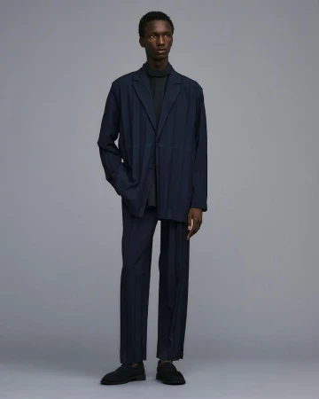 Full-length studio shot of a Black male fashion model wearing a navy suit with a dark grey turtleneck, posing against a grey background.
