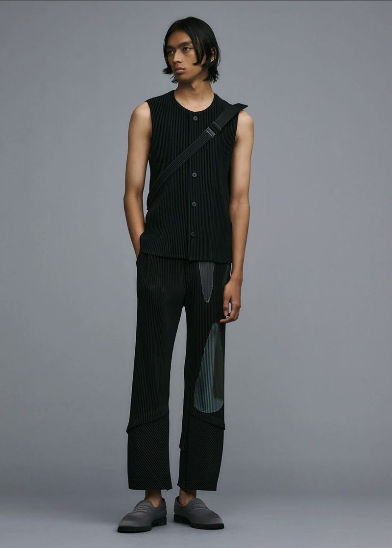 Male fashion model poses in a sleeveless black top, patch-worked trousers, and gray shoes against a solid gray backdrop.