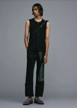 Male fashion model poses in a sleeveless black top, patch-worked trousers, and gray shoes against a solid gray backdrop.