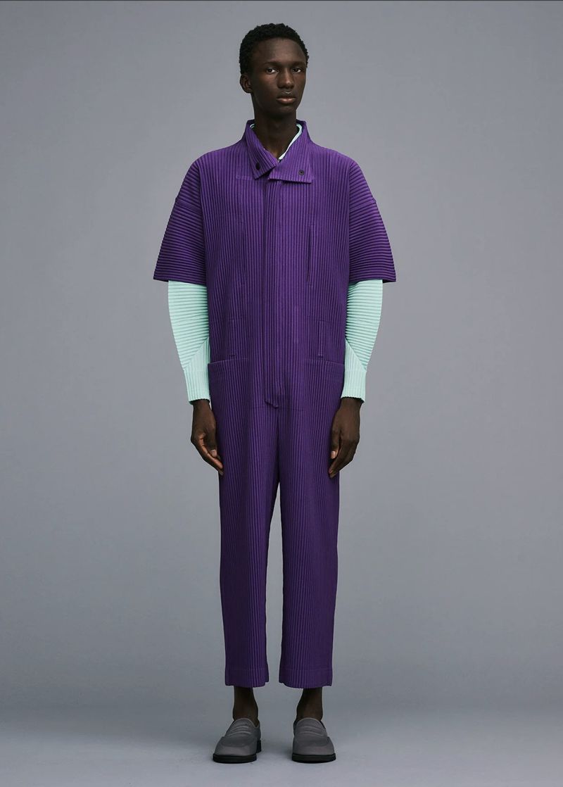 Fashion shot of a slender person wearing a purple ribbed jumpsuit with short sleeves and light blue long-sleeved shirt against gray backdrop.