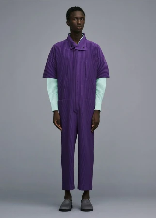 Fashion shot of a slender person wearing a purple ribbed jumpsuit with short sleeves and light blue long-sleeved shirt against gray backdrop.