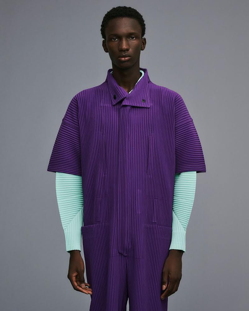A Black male fashion model poses in a pleated purple jumpsuit with mint-green long sleeves against a plain gray background.