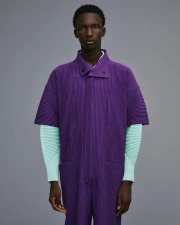 A Black male fashion model poses in a pleated purple jumpsuit with mint-green long sleeves against a plain gray background.