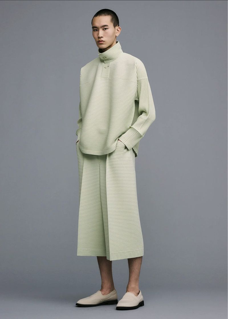 Fashionable Asian man wearing a light green pleated top and culottes with loafers stands in a studio setting. Minimalist aesthetic.