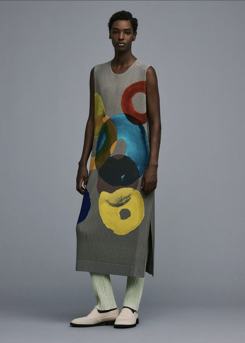 Adult female posing in grey pleated sleeveless dress with colorful circles, light-green pants, and cream loafers against a plain grey backdrop.