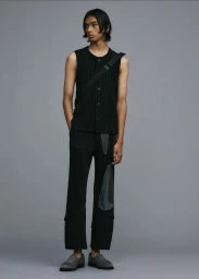 Model wearing a black pleated sleeveless top and trousers with grey loafers against a grey studio backdrop