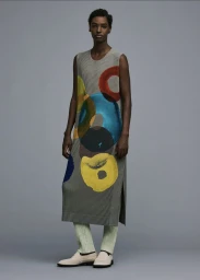 Model wearing a grey pleated tunic with colourful abstract circles and light green ribbed trousers against a plain grey background