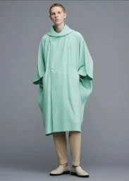 Model wearing a light green pleated coat and trousers against a grey studio backdrop