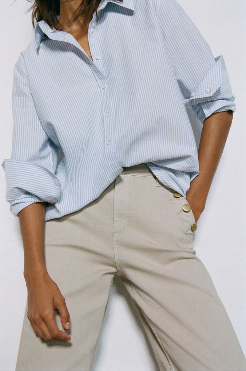 Adult female fashionably dressed in a light blue striped collared shirt and tan wide-leg pants with gold buttons, posing indoors in a studio.