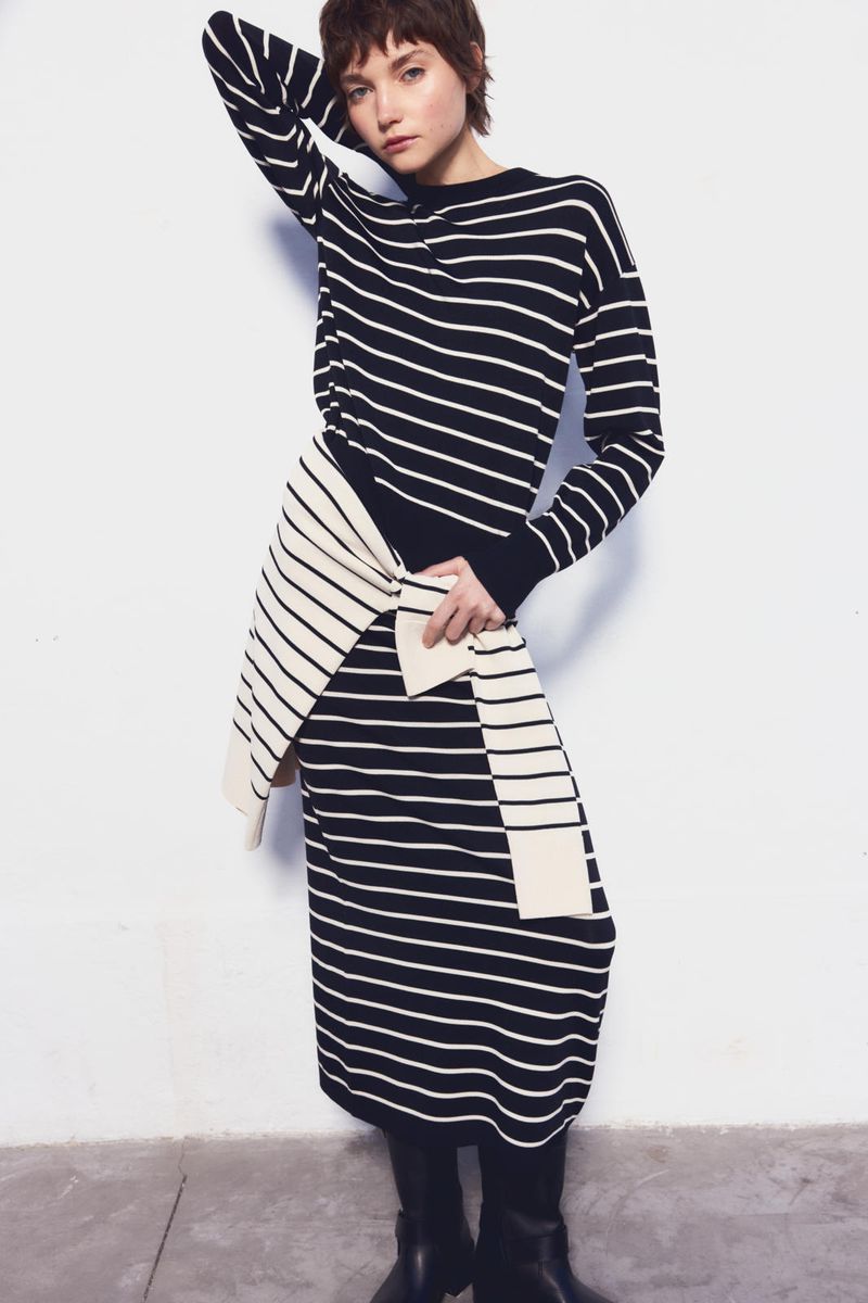 Adult female fashion model poses in black and white striped knitwear dress with long sleeves in a photo studio against a white backdrop.