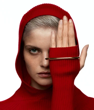 Close-up studio shot of a fair-skinned adult woman wearing a red knit hooded garment and gold bangle, partially obscuring her face.