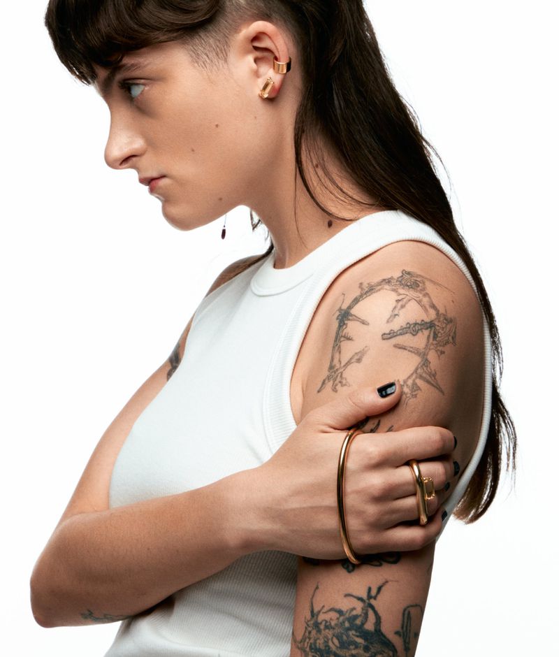 Profile of an adult woman with tattoos and jewelry against a white background, wearing a white tank top, holding out her arm.