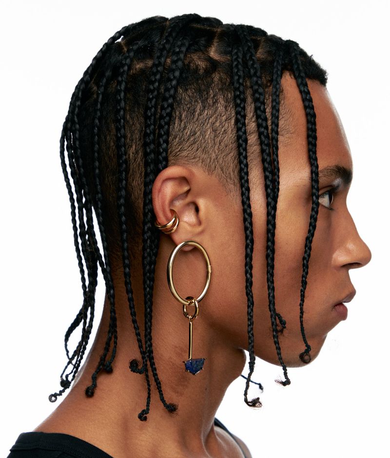 Profile portrait of a person with box braids, gold earrings, and dark clothing against a white background in a studio setting.