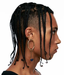 Profile portrait of a young person wearing gold hoop earrings and ear cuffs with braided black hair against a white background.