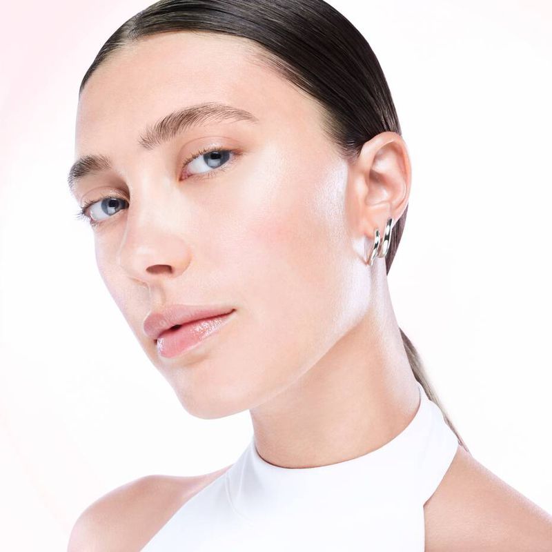 Close-up shot of a fair-skinned young adult woman with light makeup, silver hoop earrings, and a white top against a gradient background.
