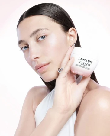 Close-up of a woman with fair skin holding Lancôme Hydra Zen Gel Cream against a white and soft pink gradient background. Beauty shot.
