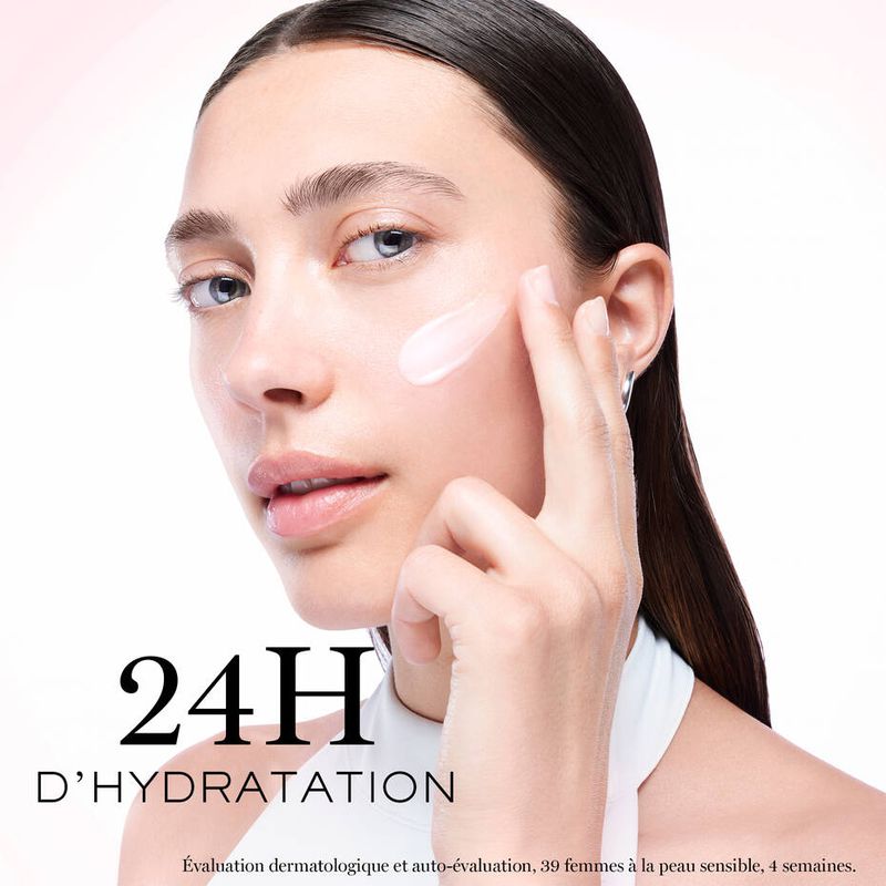 Close-up of a woman applying moisturizer to her face. She is fair-skinned with dark hair, and is in a white indoor setting.
