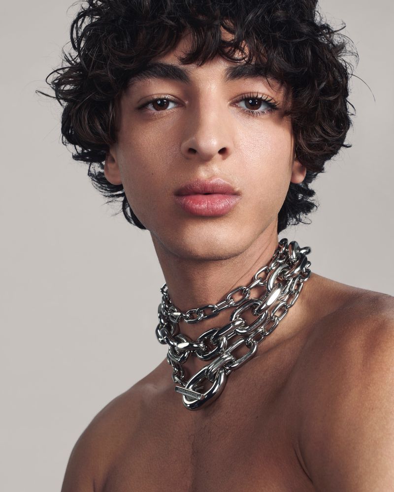 Close-up portrait of a young adult male with curly hair and a heavy silver chain necklace on a neutral background in studio lighting.