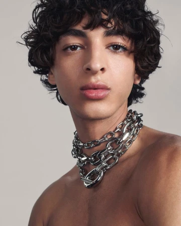 Close-up portrait of a young adult male with curly hair and a heavy silver chain necklace on a neutral background in studio lighting.