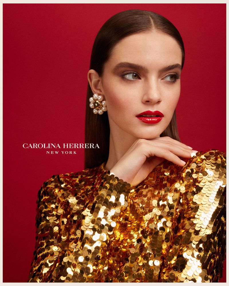 Close-up of a woman with dark hair, wearing a gold sequin top and large pearl earrings, against a red background for a fashion ad.
