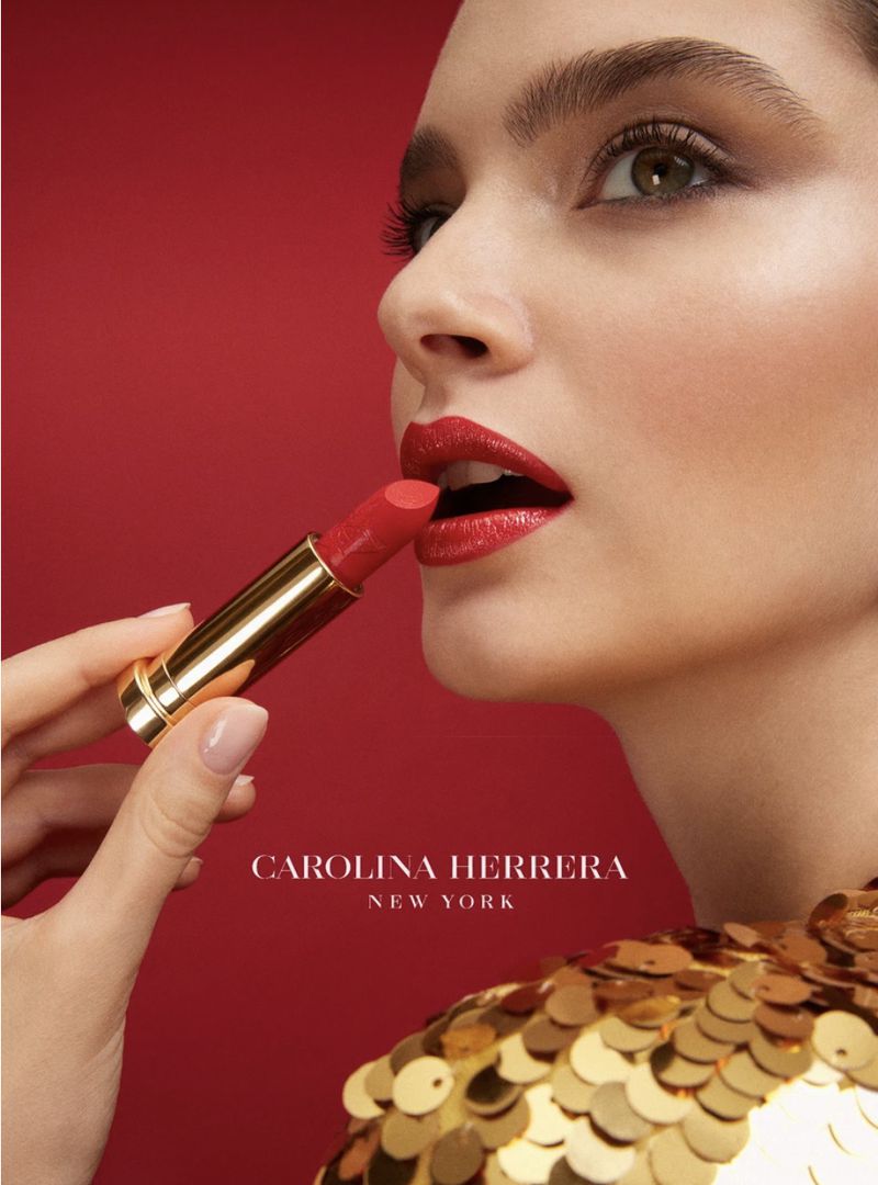 Glamorous beauty shot of a woman with red lipstick, holding the tube in her hand against a red background, wearing a gold sequin top.