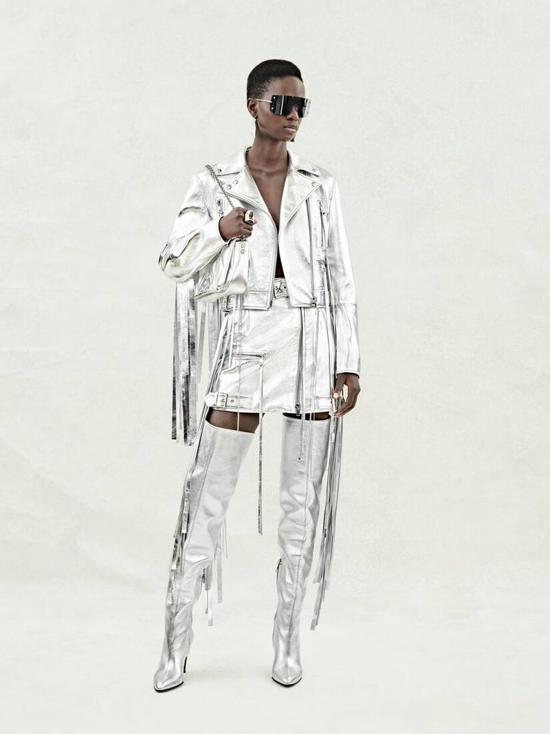 A slender Black female model poses in a shiny silver motorcycle jacket, mini-skirt, thigh-high boots, and dark sunglasses against a plain background.