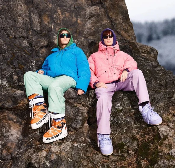 Two people in bright outerwear sitting on a rock outcrop with a blurred treeline background, stylishly dressed with eye-catching footwear.