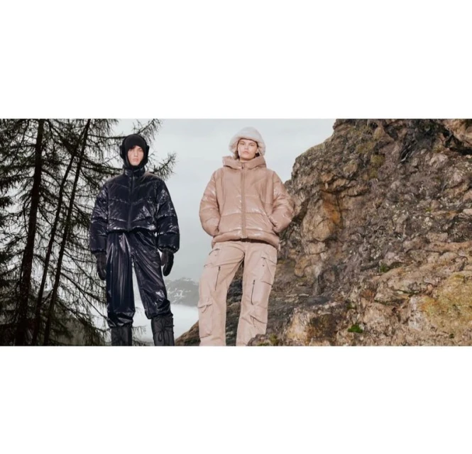 Two people in matching winter outfits stand on a rocky outcrop in a cold environment with evergreen trees and distant snowy mountains.