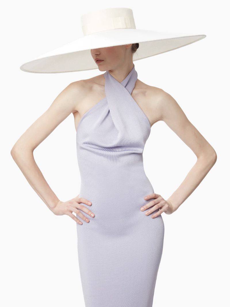 Elegant woman posing in a lilac dress and a wide-brimmed white hat against a white backdrop in a studio setting, showcasing fashion and style.