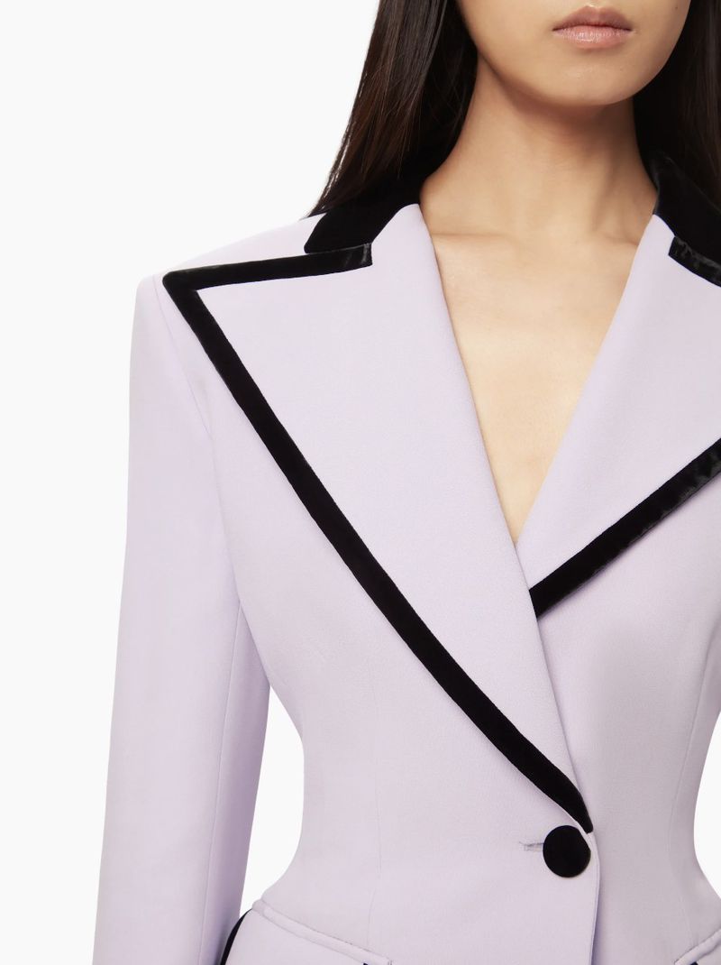 Close-up shot of a woman's upper torso wearing a light purple blazer with black trim, emphasizing fashion and detail against a white backdrop.