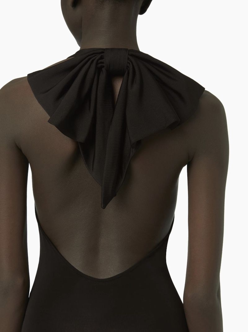 Back view of an African American woman in a black dress with a large bow at the neck, set against a plain white background.