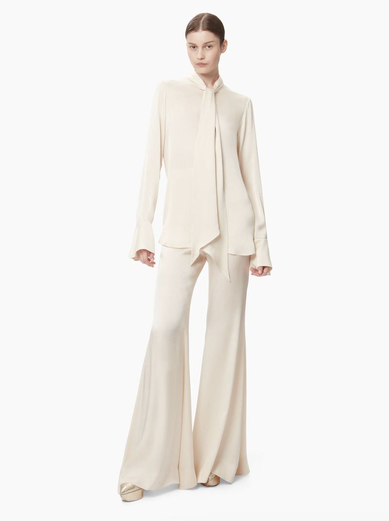 Elegant adult woman in studio wearing flowing ivory blouse with bow and flared pants in minimalist fashion pose on white background.