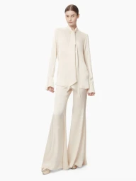 Full-length studio portrait of a female model wearing a cream silk blouse with a bow tie collar and matching flared trousers against a white background.
