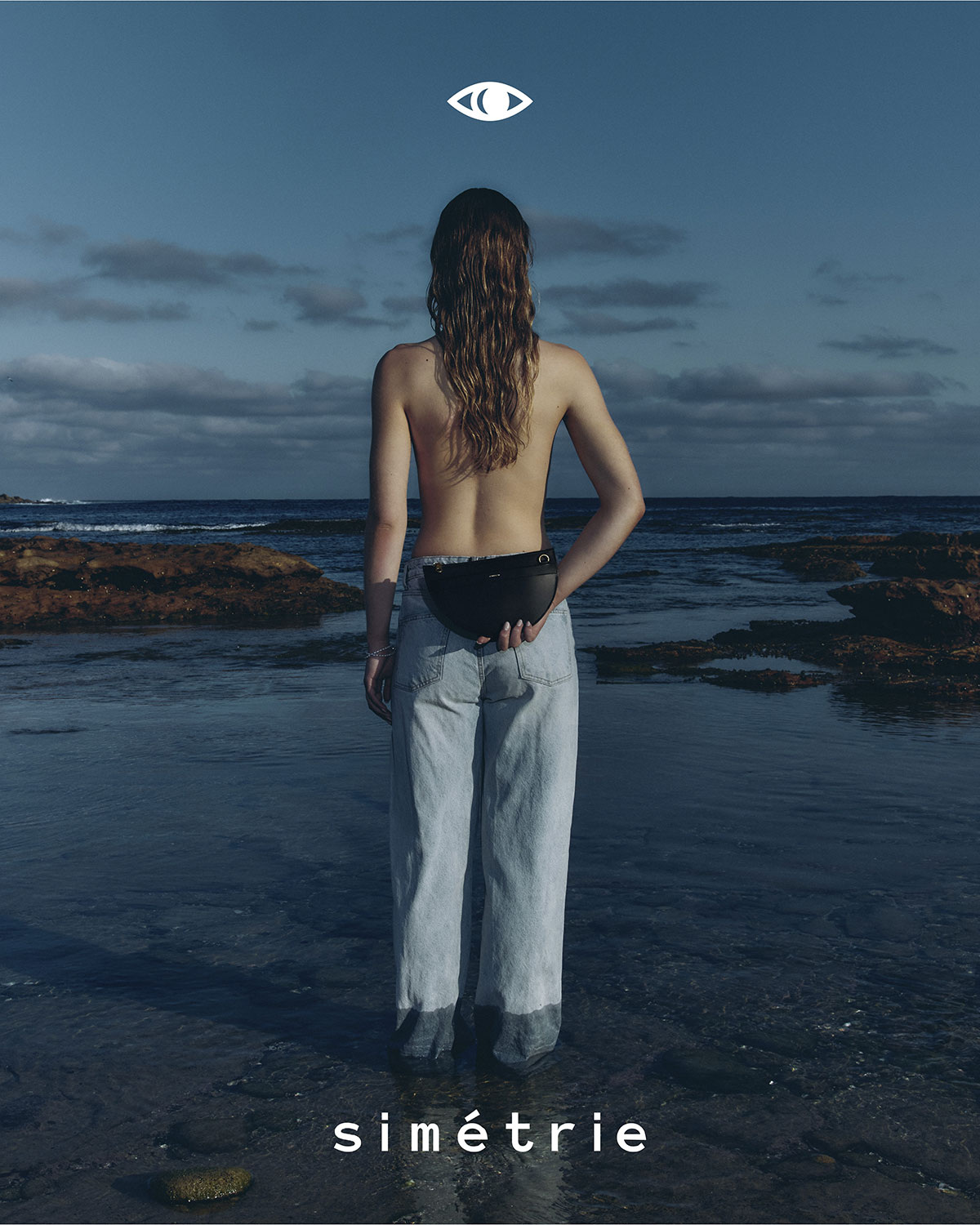 Woman stands in ocean water facing away from camera, wearing jeans and black bag, under eye-like graphic on blue cloudy sky backdrop.
