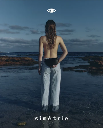 Woman stands in ocean water facing away from camera, wearing jeans and black bag, under eye-like graphic on blue cloudy sky backdrop.