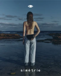Model seen from behind wearing light-wash denim jeans and holding a black leather handbag against a rocky coastal backdrop.