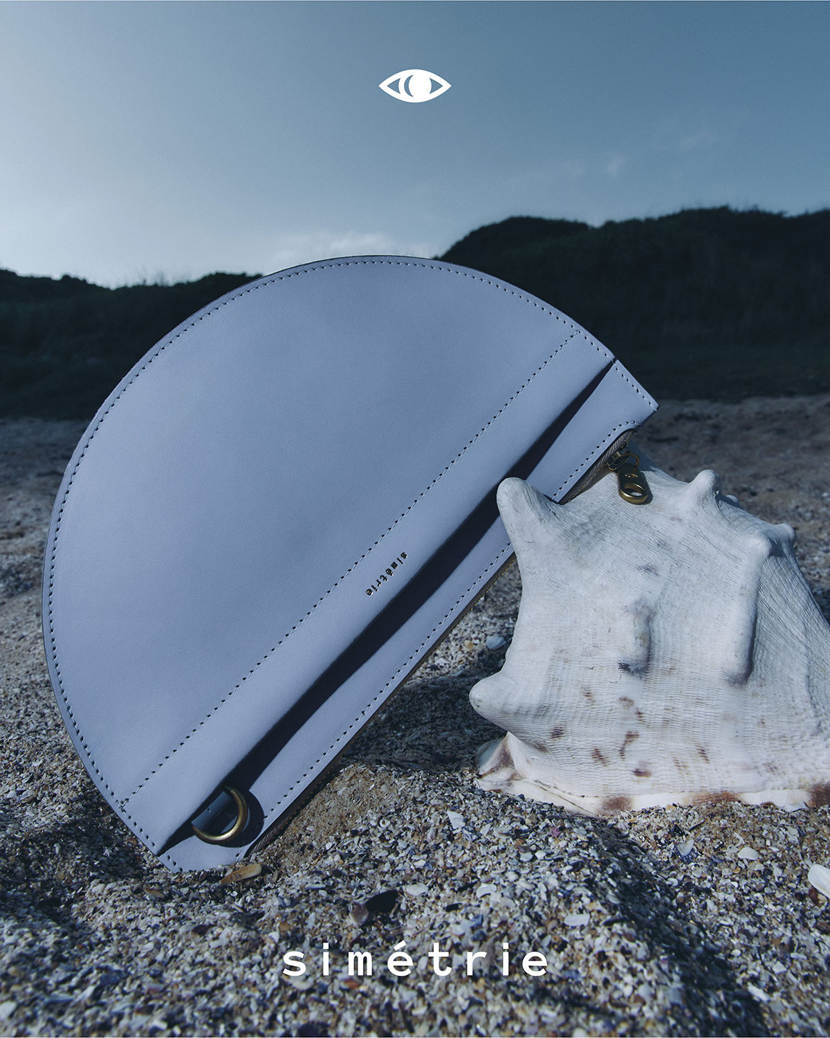 Fashion shot of a light blue Simetrie handbag on a beach, next to a large shell, under an overcast sky with the brand logo.