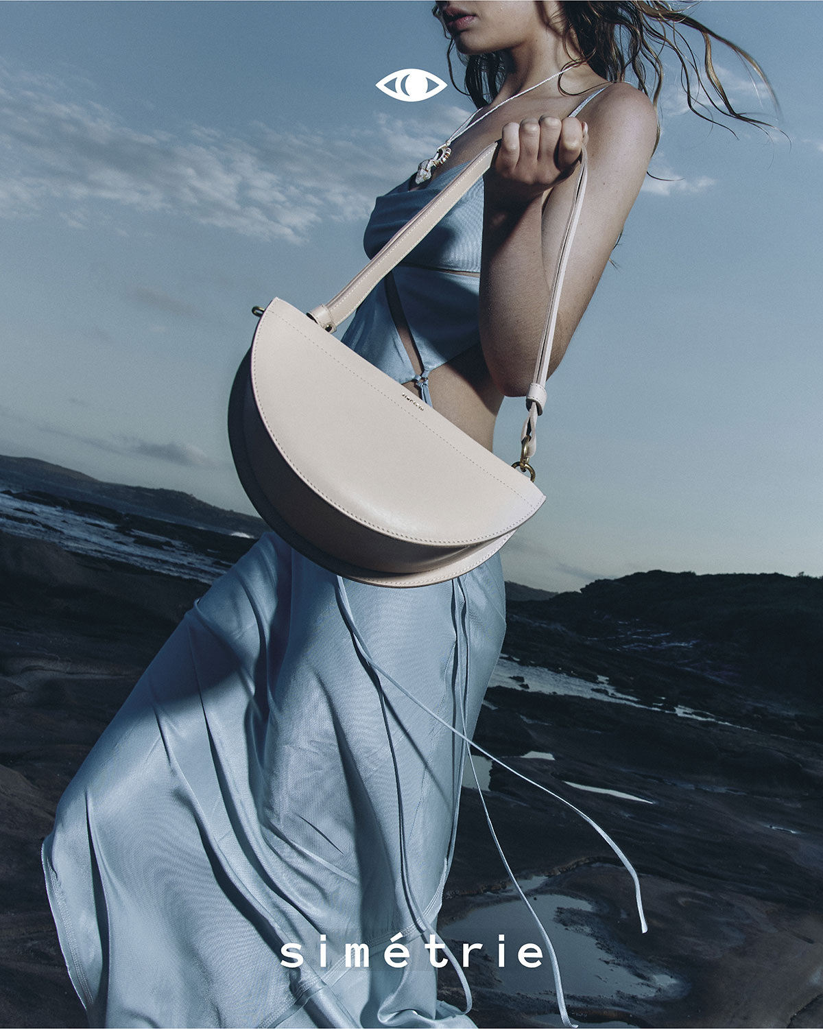 An adult female fashion model poses outdoors with a cream-colored, half-moon shaped bag with thin straps, in front of an ocean and rock formation.