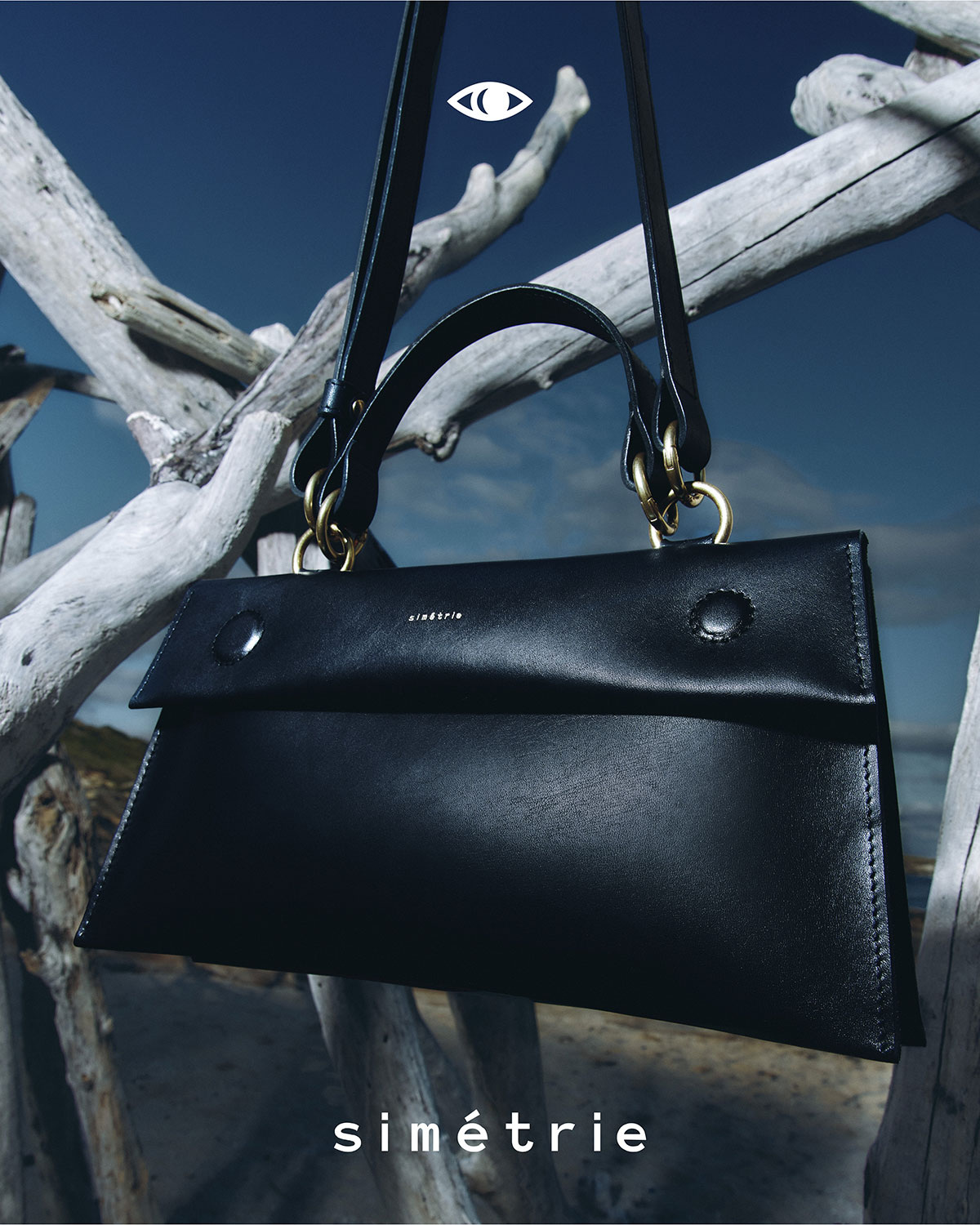 Elevated product shot of a black leather handbag with gold accents hanging from driftwood against a blue sky for a fashion advertisement.