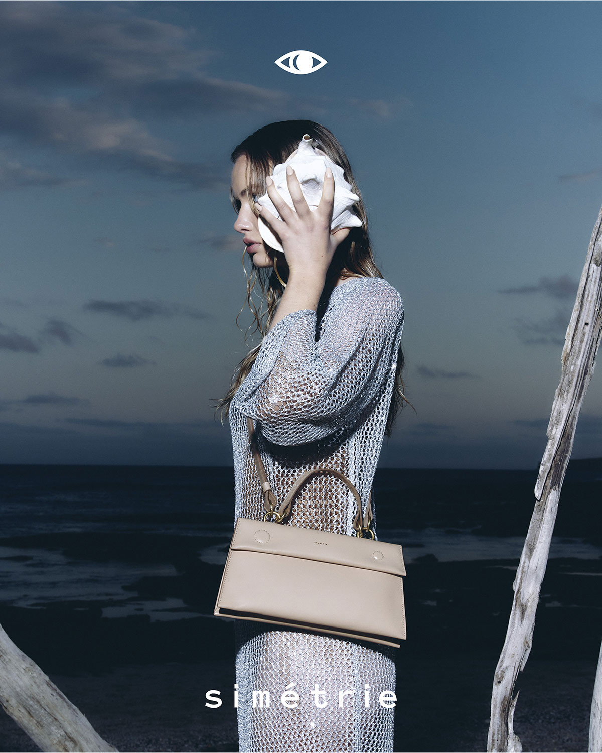 Model holding a large white shell to her ear, wearing a sheer metallic dress and carrying a beige handbag outdoors near water.