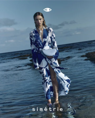 Woman in a blue and white floral-patterned dress stands on a rocky beach with a blue sky backdrop, wearing a necklace and handbag.