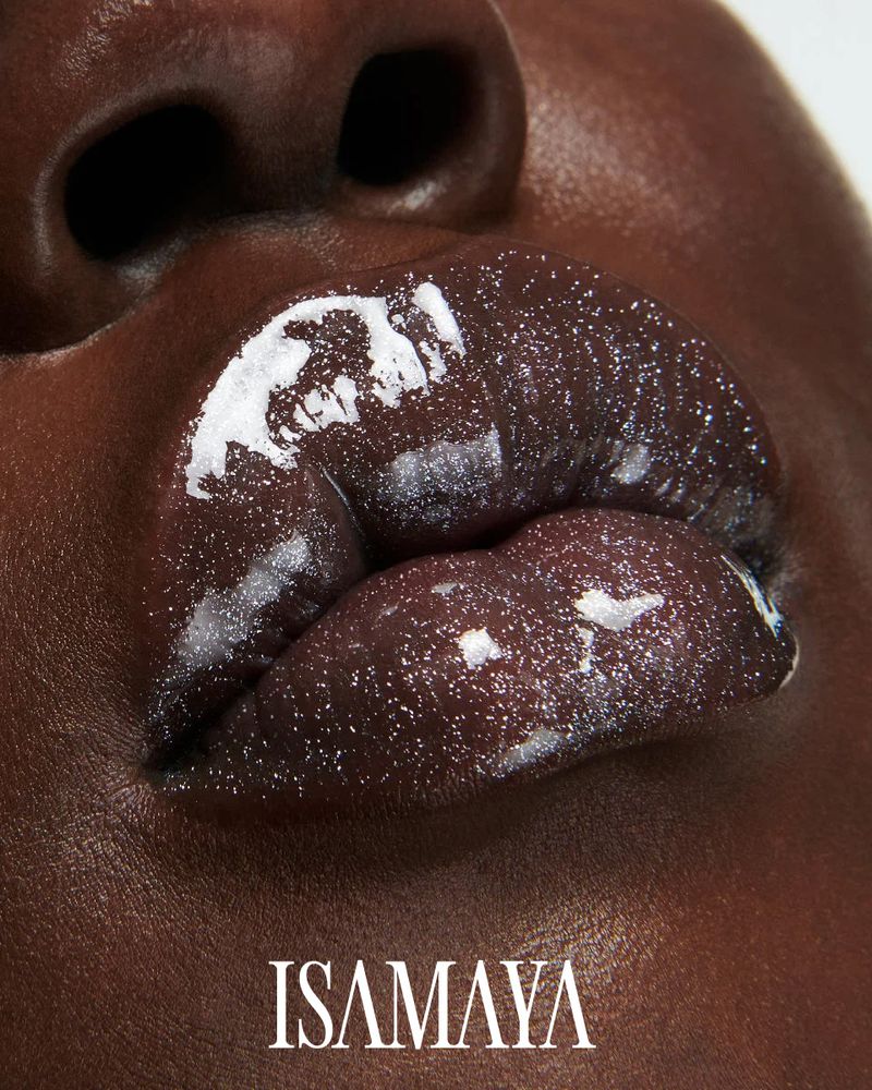 Close-up shot of a dark-skinned adult female showcasing glossy, dark-colored lip makeup and the brand logo 'ISAMAYA' in the foreground.